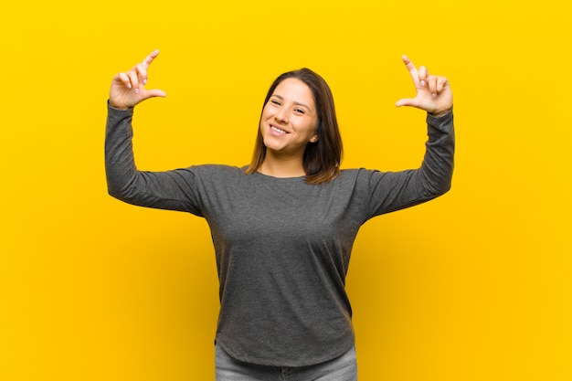 Latynoamerykańska kobieta obramia lub obrysowywa własnego uśmiech obiema rękami, patrzeje pozytywny i szczęśliwy, wellness pojęcie odizolowywający przeciw kolor żółty ścianie