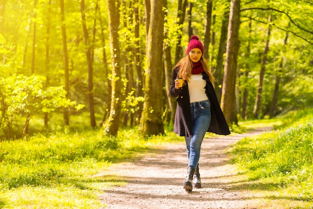Latynoska dziewczyna w czarnej kurtce, szaliku i czerwonym wełnianym kapeluszu, ciesząca się jesiennym parkiem, idąca ścieżką z kawą o zachodzie słońca