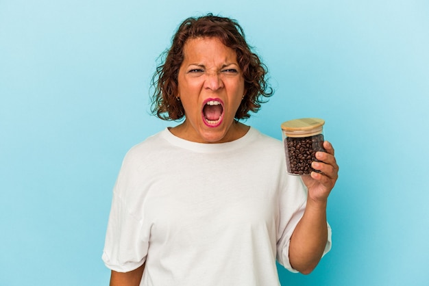 Latynoska kobieta w średnim wieku trzymająca słoik kawy na białym tle na niebieskim tle krzyczy bardzo zła i agresywna.