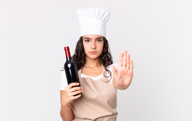 Latynoska ładna szefowa kuchni wygląda poważnie, pokazując otwartą dłoń, wykonując gest zatrzymania i trzymając butelkę wina