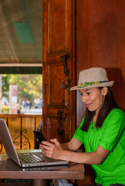 Latynoska w kapeluszu i zielonej bluzce siedzi w kawiarni, sprawdzając smartfon i pracując na laptopie