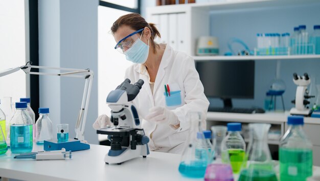 Latynoska w średnim wieku ubrana w mundur naukowca i maskę medyczną przy użyciu mikroskopu w laboratorium