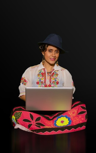 Latynoska w tradycyjnej sukience i kapeluszu siedzi na podłodze, trzymając laptopa na nogach, uśmiechając się w ciemnym tle