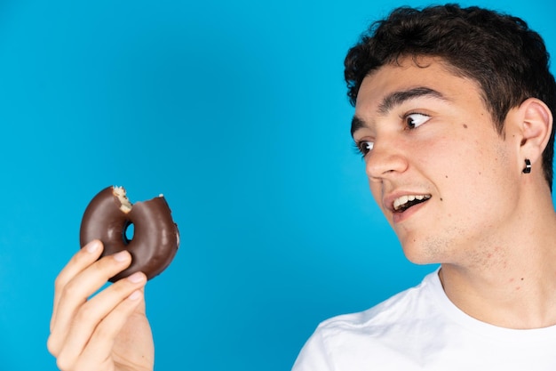 Latynoski nastolatek chłopiec patrzący na czekoladowego pączka lub pączka z zazdrością na białym tle na niebieskim tle