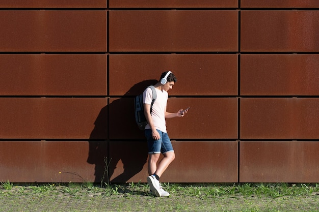 Latynoski student słuchający muzyki i sprawdzający swój telefon oparty o ścianę miasta