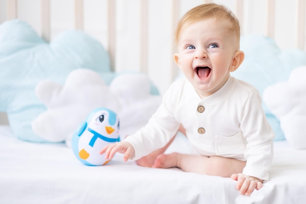 Laughing baby 6 miesięcy blond chłopiec w łóżeczku w jasnej sypialni w białym bawełnianym body z niebieską zabawką koncepcja artykułów dziecięcych