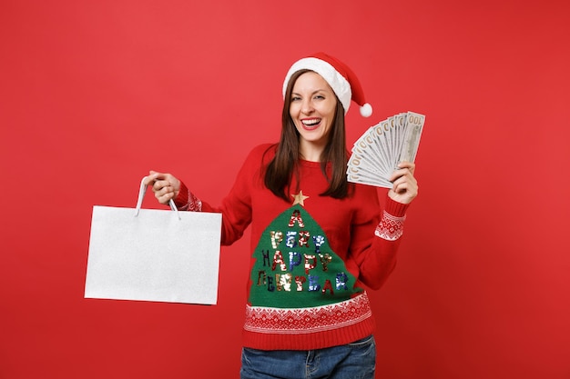 Lauging młoda dziewczyna Santa trzymać wiele banknotów dolarów, gotówki pakiety pieniędzy torby z zakupami po zakupach na białym tle na czerwonym tle. Szczęśliwego nowego roku 2019 celebracja party wakacje koncepcja.