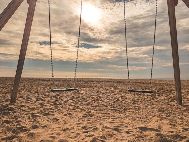 Ławki wiszące na drewnianych słupkach na piaszczystej plaży z łańcuchami w słonecznym świetle przeciwko chmurom