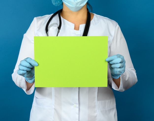 Zdjęcie lekarka w białym fartuchu medycznym, jednorazowej masce i czapce stoi na niebieskiej ścianie i trzyma pustą zieloną kartkę papieru, miejsce na napis