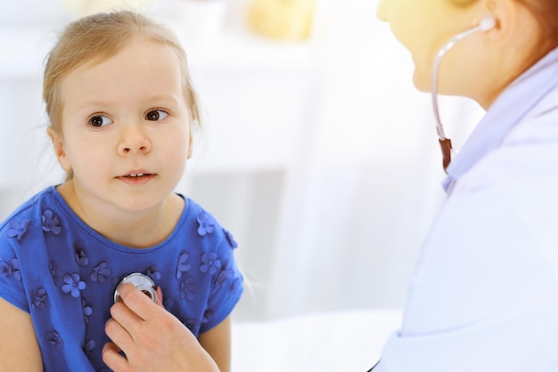 Lekarz bada małą dziewczynkę za pomocą stetoskopu. Szczęśliwy uśmiechnięty dziecięcy pacjent podczas zwykłej inspekcji medycznej. Koncepcje medycyny i opieki zdrowotnej.