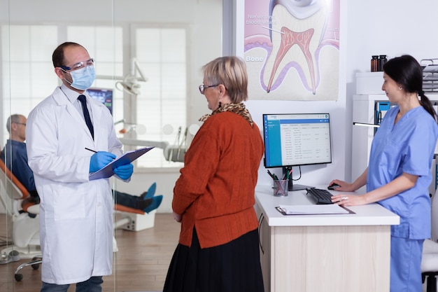 Lekarz dentysta z maską wyjaśniającą diagnozę pacjentowi starszej kobiety w korytarzu poczekalni stomatologii. Starszy mężczyzna siedzi na krześle do leczenia zębów.