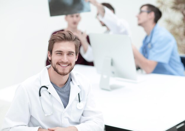 Lekarz patrzący w kamerę i uśmiechający się, podczas gdy jego koledzy siedzą w tle
