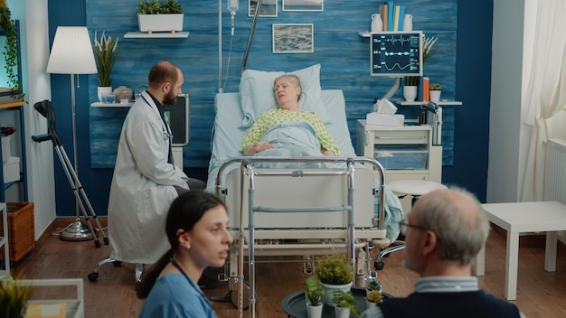 Lekarz rozmawia z pacjentem w wieku z kroplówką IV w domu opieki, leżąc na łóżku szpitalnym. Medyk udzielający pomocy chorej na emeryturze kobiecie w leczeniu i lekach