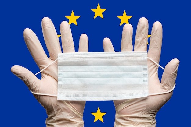 Lekarz trzymający maskę medyczną w dwóch rękach w białych rękawiczkach na tle flagi Unii Europejskiej UE