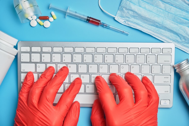 Lekarz w czerwonych rękawiczkach pracuje przy komputerze na niebieskim tle w pobliżu sprzętu medycznego.