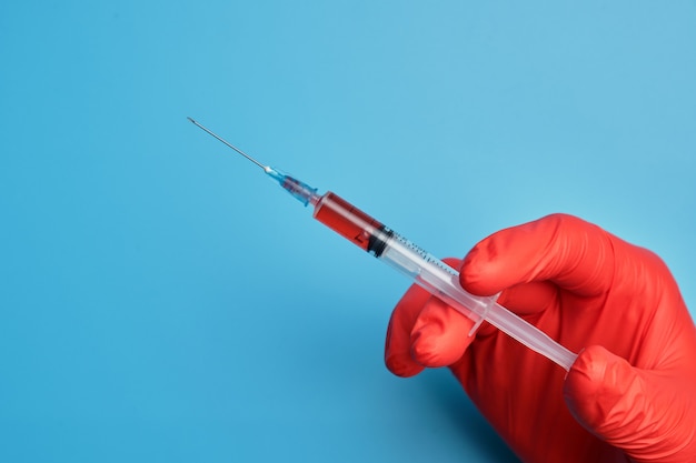 Lekarz w czerwonych rękawiczkach trzyma strzykawkę ze szczepionką na niebieskim tle.