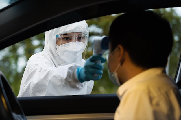 Lekarz w ochronnym kombinezonie PPE za pomocą termometru na podczerwień mierzącego temperaturę człowieka na stacji drive thru, IR Thermoscan