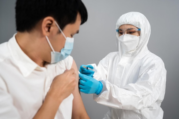 Lekarz w strzykawce ochronnego kombinezonu PPE i za pomocą bawełny przed wykonaniem wstrzyknięcia pacjentowi w masce medycznej. Szczepionka przeciw Covid-19 lub koronawirusowi