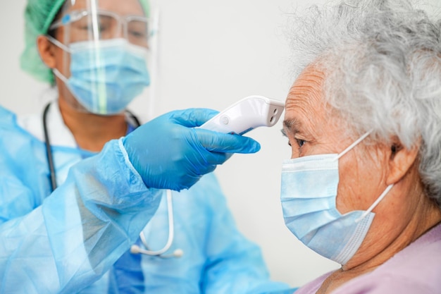 Lekarz w termometrze na podczerwień użytkownika PPE, aby sprawdzić temperaturę pacjenta w podeszłym wieku azjatyckiej kobiety dla bezpieczeństwa w celu ochrony wirusa Covid 19