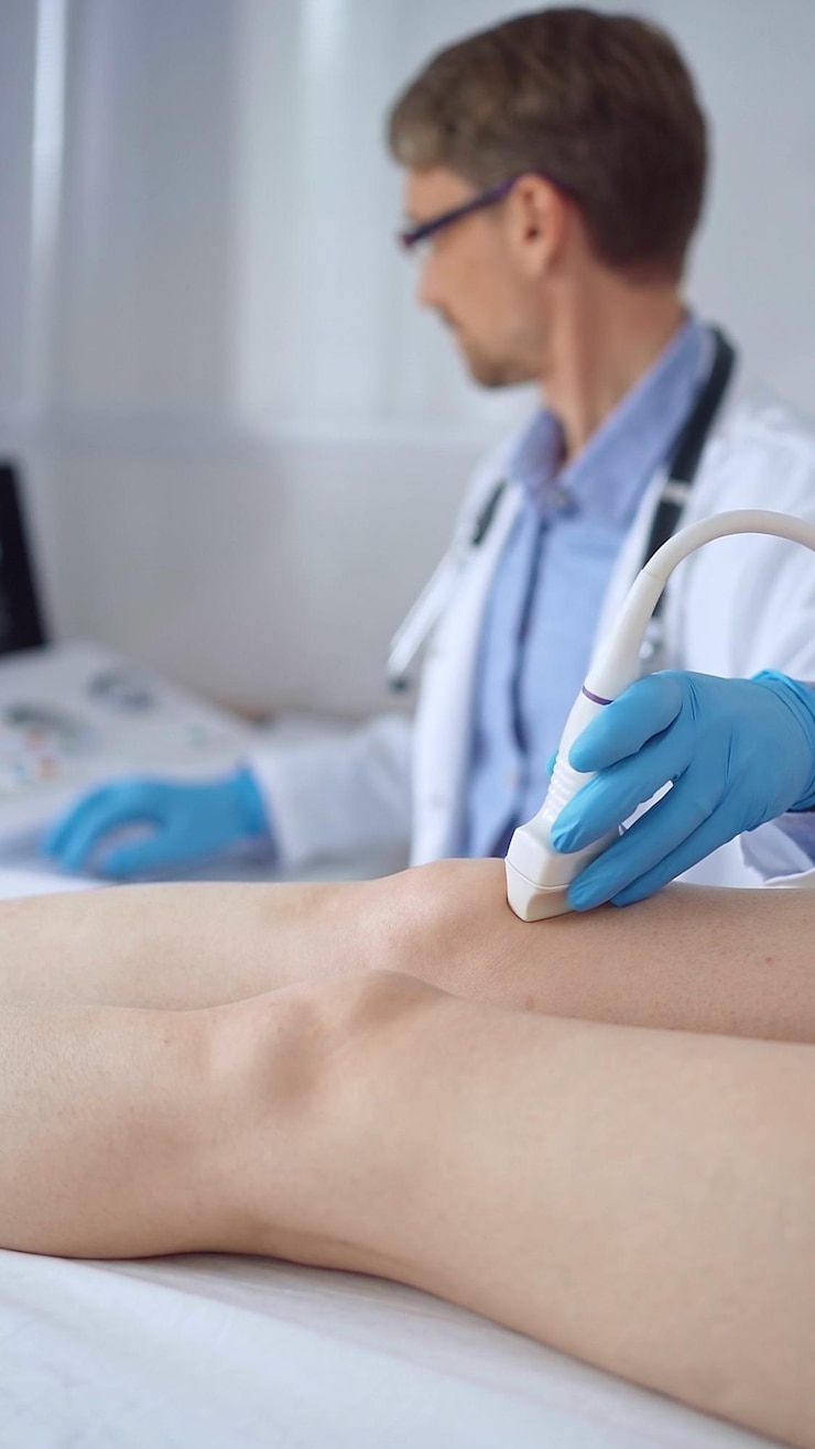 Zdjęcie lekarz z niebieskimi rękawiczkami ochronnymi używający sprzętu ultradźwiękowego na kolanie pacjentki analizujący wyniki na monitorze w tle medycyna