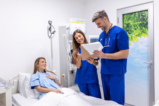 Lekarze w mundurach ze stetoskopem używający schowka stojący na oddziale szpitalnym z pacjentem odpoczywającym w łóżku na tle Lekarz sprawdzający dane pacjenta
