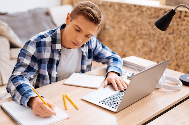 Lekcje Uczenia Się. Atrakcyjny Poważny Jasnowłosy Chłopak Pracuje Na Swoim Laptopie I Pisze W Swoim Notesie, Siedząc Przy Stole