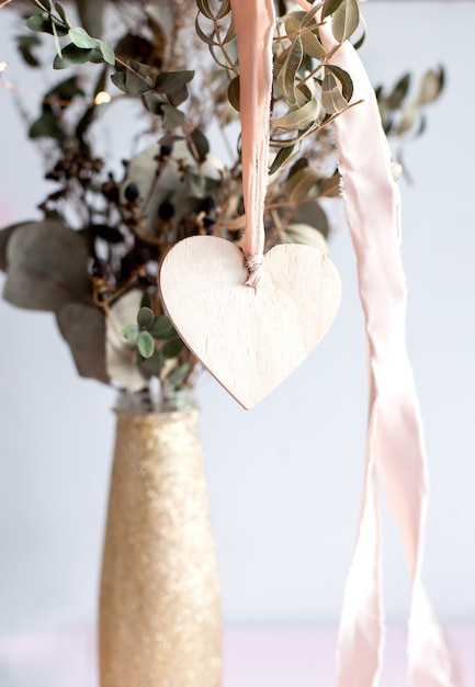 Zdjęcie lekka drewniana zawieszka w kształcie serca na różowej jedwabnej wstążce na tle błyszczącego złotego wazonu z suszonymi gałązkami eukaliptusa.