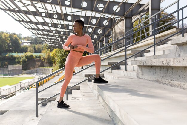 Lekkoatletka uprawia fitness w różowym stroju sportowym w pobliżu stadionu sportowego, Afroamerykanka wykonuje ćwiczenia fitness i rozciąga się rano
