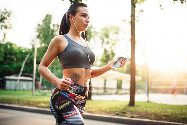 Lekkoatletyczna dziewczyna jogging ze słuchawkami na chodniku w parku latem. Kobieta na zewnątrz rano treningu