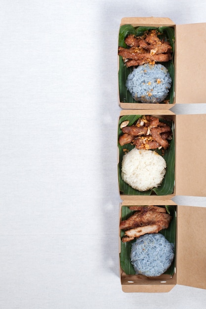 Lepki ryż z grillowaną wieprzowiną i smażoną wieprzowiną włóż w brązowe papierowe pudełko, połóż na białym obrusie, pudełku na żywność, tajskie jedzenie.