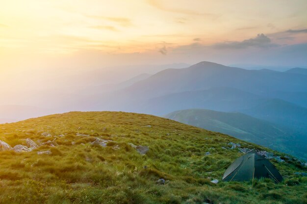 Letni Camping W Górach O świcie. Namiot Turystyczny Na Okrągłym Trawiastym Wzgórzu Na Odległych Górach Mglistych Niebieskich Gór Pod Różowym Niebem Przed Wschodem Lub Zachodem Słońca. Pojęcie Turystyki, Turystyki Pieszej I Piękna Przyrody.