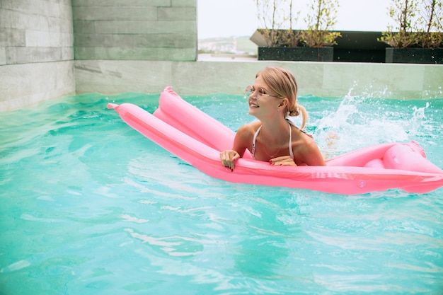 Letni obraz oszałamiającej opalonej dziewczyny pływającej z różowym materacem w basenie i śmiejącej się. Ma na sobie stylowe białe bikini i różowe okulary przeciwsłoneczne. Uśmiechać się i cieszyć życiem. Opalanie się