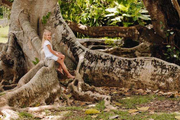 Letni portret szczęśliwej małej dziewczynki na wyspie Mauritius siedzącej na ogromnym drzewie. Dziewczyna siedzi na dużym drzewie w ogrodzie botanicznym na wyspie Mauritius