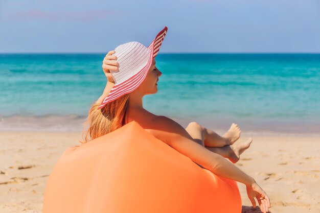 Letni styl życia portret ładna dziewczyna siedzi na pomarańczowej nadmuchiwanej sofie na plaży tropikalnej wyspie, relaksując się i ciesząc się życiem na łóżku powietrznym