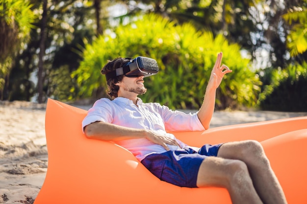 Letni styl życia portret mężczyzny siedzącego na pomarańczowej nadmuchiwanej sofie i korzysta z zestawu słuchawkowego wirtualnej rzeczywistości na plaży na tropikalnej wyspie Relaksujący i cieszący się życiem na dmuchanym łóżku