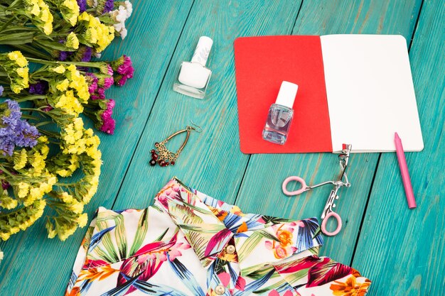 Letni zestaw damski z sukienką kolorowe kwiaty kosmetyki do makijażu notatnik bijou i podstawowe produkty na niebieskim drewnianym tle