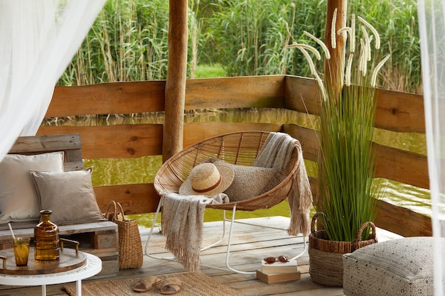 letnia altana nad jeziorem ze stylowym fotelem rattanowym, stolikiem kawowym, sofą, poduszkami, kratą i eleganckimi dodatkami w nowoczesnym wystroju.