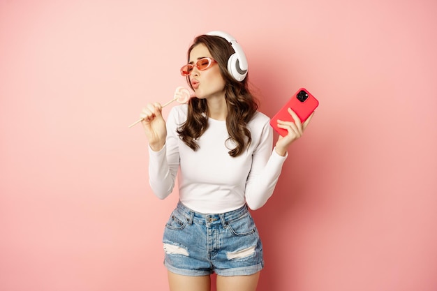 Letnia dziewczyna śmiejąc się, jedząc lolipop i słuchając muzyki w słuchawkach, tańcząc ze smartfonem na różowym tle.