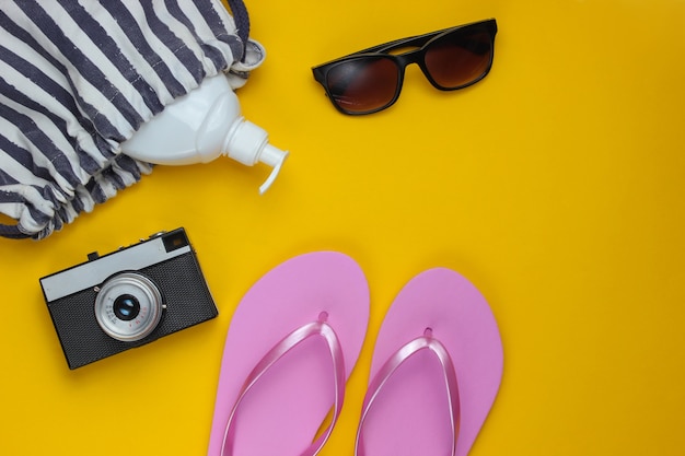 Letnia martwa natura. Akcesoria plażowe. Modne różowe klapki, torba, aparat retro, butelka z filtrem, okulary przeciwsłoneczne, muszla na żółtym tle papieru.