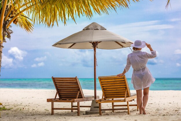 Letnia młoda kobieta stojąca do tyłu i oglądająca plażę w białej sukni, kapeluszu. Letnia plaża relaksacyjna