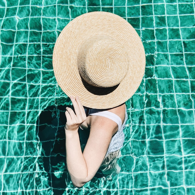 Letnia podróż koncepcja mody i wakacji. Piękna młoda, opalona kobieta w słomkowym kapeluszu i białym kostiumie kąpielowym stoi przy basenie