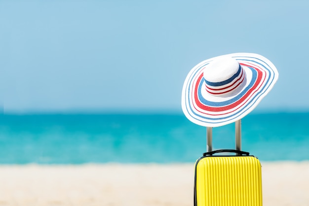 Letnie podróże i planowanie z żółtym bagażem walizkowym z modnym kapeluszem na piaszczystej plaży. Podróżuj na tle wakacji, niebieskiego nieba i plaży. Koncepcja lato i podróż
