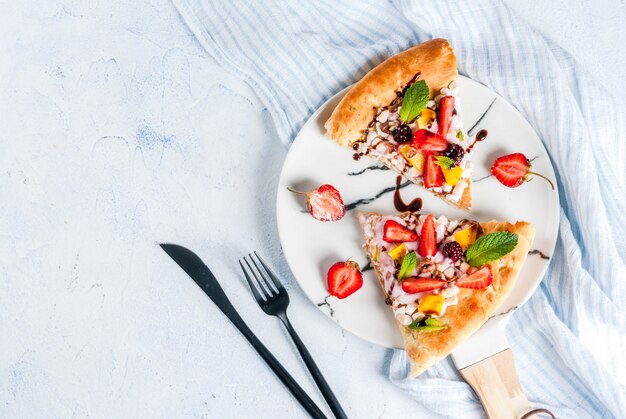 Letnie przekąski. Jedzenie na imprezę. Pizza owocowa ze śmietaną, porzeczkami, jogurtem, truskawkami, mango, brzoskwiniami, bananami, jeżynami, czekoladą, orzechami włoskimi, miętą. Na jasnoniebieskim stole. widok z góry lato