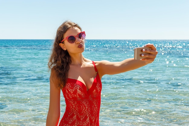 Letnie wakacje na plaży dziewczyna robiąca zabawne mobilne zdjęcie selfie ze smartfonem Dziewczyna w czerwonych okularach przeciwsłonecznych pozuje do selfie