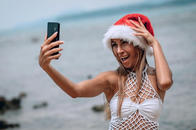 Letnie wakacje na plaży dziewczyna w santa hat biorąc zabawne zdjęcie mobilne selfie z smartphone. Dziewczyna ubrana w biały strój kąpielowy pozowanie do selfie. Wyjazdy do ciepłych miejsc. Phuket. Tajlandia.