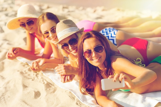 Zdjęcie letnie wakacje, podróże, technologia i koncepcja ludzi - grupa uśmiechniętych kobiet w okularach przeciwsłonecznych i kapeluszach robi selfie ze smartfonem na plaży
