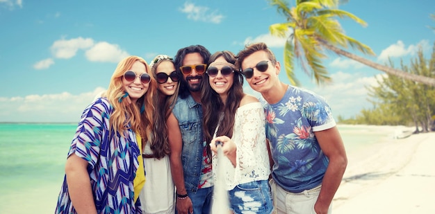 letnie wakacje, podróże, turystyka, technologia i koncepcja ludzi - uśmiechnięci młodzi przyjaciele hipisów, którzy robią zdjęcie za pomocą smartfona selfie stick na tle plaży