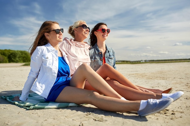 letnie wakacje, wakacje, podróże i koncepcja ludzi - grupa uśmiechniętych młodych kobiet w okularach przeciwsłonecznych i codziennych ubraniach na opalaniu się na kocu plażowym