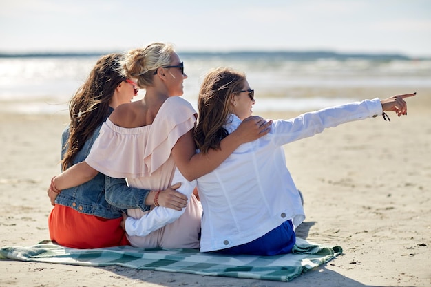 Zdjęcie letnie wakacje, wakacje, podróże i koncepcja ludzi - grupa uśmiechniętych młodych kobiet w okularach przeciwsłonecznych siedzących na kocu plażowym i wskazujących na coś palcem