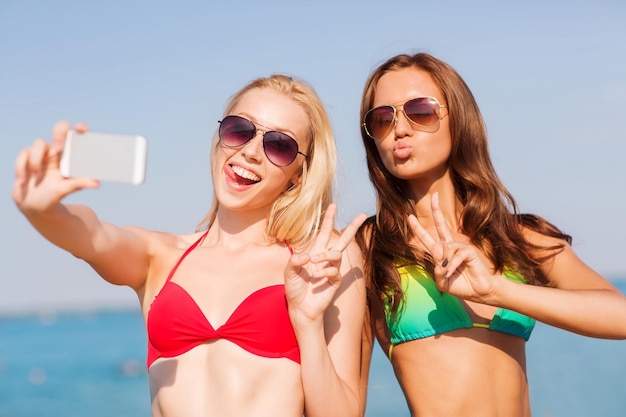 letnie wakacje, wakacje, podróże, technologia i koncepcja ludzi - dwie uśmiechnięte młode kobiety na plaży robiące selfie ze smartfonem na tle błękitnego nieba